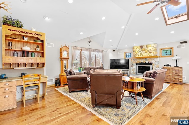 living area featuring ceiling fan, vaulted ceiling with skylight, a stone fireplace, light wood-type flooring, and built in desk