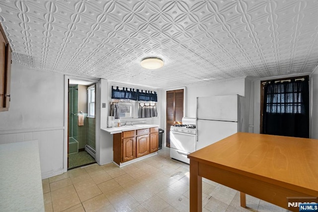 kitchen with white appliances, brown cabinetry, wainscoting, baseboard heating, and light countertops