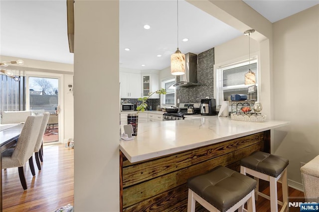 kitchen featuring white cabinets, wall chimney exhaust hood, glass insert cabinets, light countertops, and pendant lighting