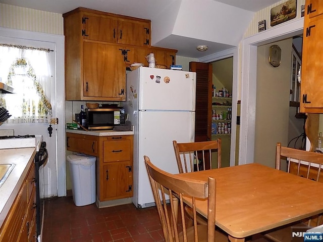 kitchen featuring stove, brown cabinets, freestanding refrigerator, brick floor, and light countertops