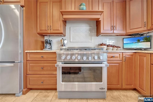 kitchen featuring light tile patterned floors, stainless steel appliances, premium range hood, brown cabinets, and tasteful backsplash