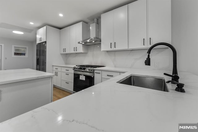 kitchen featuring stainless steel gas stove, wall chimney exhaust hood, light stone countertops, black refrigerator with ice dispenser, and a sink