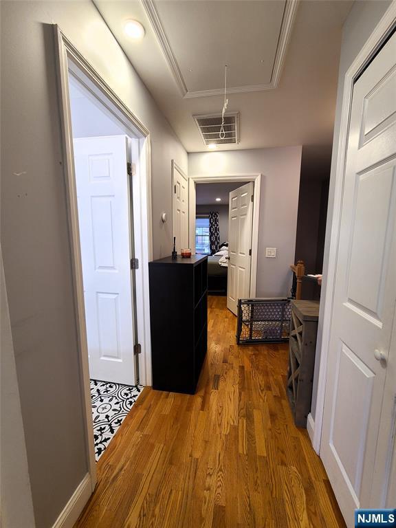 corridor with a raised ceiling, visible vents, attic access, ornamental molding, and wood finished floors