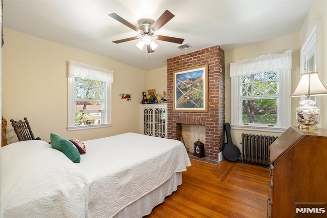 bedroom featuring ceiling fan, visible vents, a brick fireplace, wood finished floors, and radiator heating unit