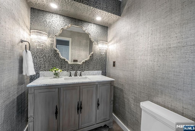 half bath with baseboards, vanity, toilet, and wallpapered walls