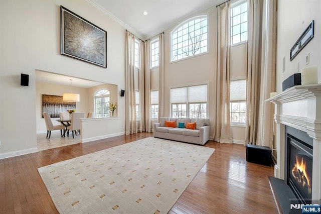 living room with baseboards, a glass covered fireplace, wood-type flooring, a high ceiling, and crown molding