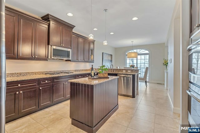 kitchen with dark brown cabinetry, appliances with stainless steel finishes, light stone countertops, an island with sink, and pendant lighting