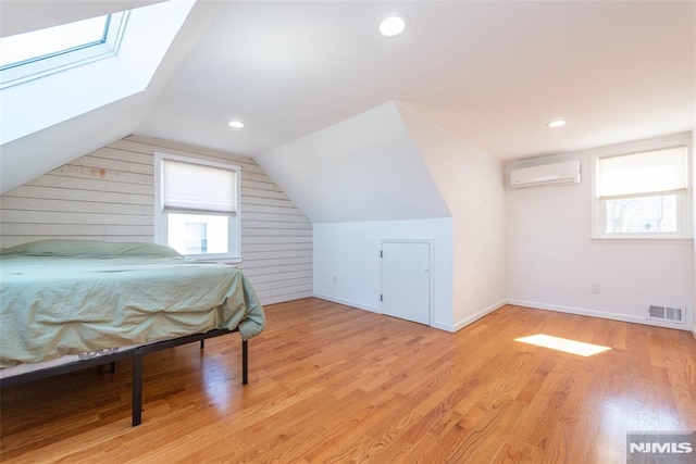 bedroom featuring visible vents, a wall mounted AC, light wood-type flooring, vaulted ceiling with skylight, and baseboards