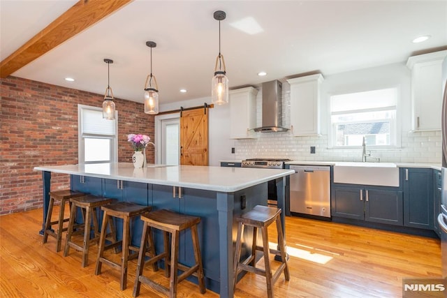 kitchen with a barn door, appliances with stainless steel finishes, a sink, brick wall, and wall chimney exhaust hood