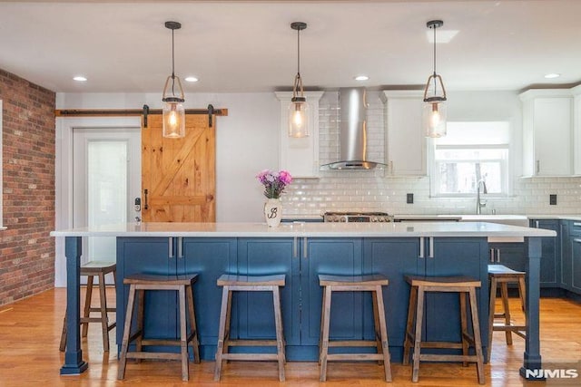 kitchen with a barn door, light wood-style floors, brick wall, light countertops, and wall chimney range hood