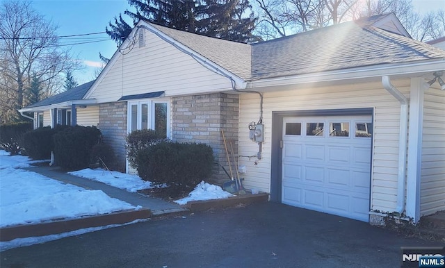 snow covered property with a garage, stone siding, and roof with shingles