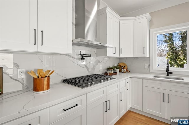 kitchen with tasteful backsplash, stainless steel gas stovetop, white cabinets, a sink, and wall chimney range hood