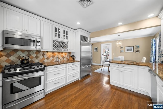 kitchen with visible vents, appliances with stainless steel finishes, glass insert cabinets, dark stone countertops, and decorative light fixtures