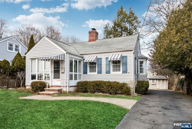 view of front of house featuring a shingled roof, a chimney, an outbuilding, a shed, and a front yard