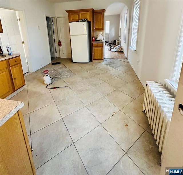 kitchen with arched walkways, light countertops, freestanding refrigerator, radiator, and brown cabinetry