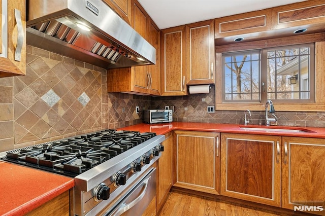 kitchen with high end stove, a sink, exhaust hood, brown cabinetry, and dark countertops