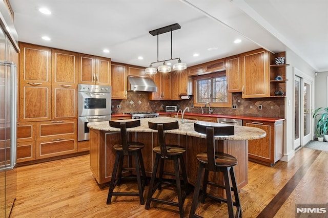 kitchen with stainless steel appliances, wall chimney range hood, open shelves, an island with sink, and decorative light fixtures