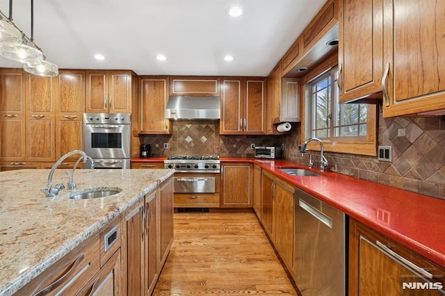 kitchen with a sink, appliances with stainless steel finishes, range hood, brown cabinetry, and pendant lighting