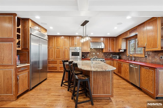 kitchen with light stone counters, pendant lighting, stainless steel appliances, a kitchen island with sink, and wall chimney exhaust hood