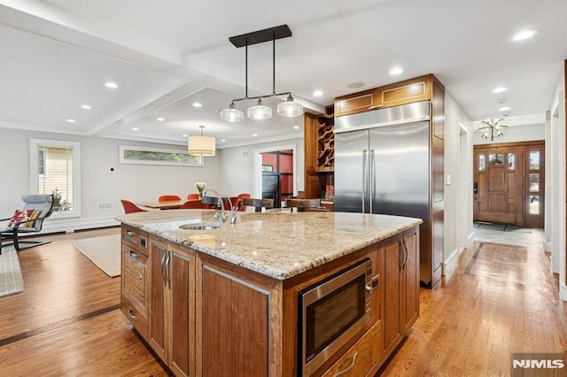 kitchen with brown cabinetry, an island with sink, decorative light fixtures, built in appliances, and light stone countertops