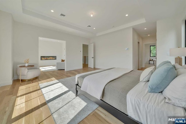 bedroom featuring a tray ceiling, recessed lighting, visible vents, light wood-style floors, and a glass covered fireplace