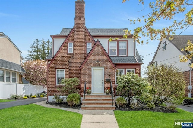 tudor home with roof with shingles, brick siding, fence, a front lawn, and stucco siding