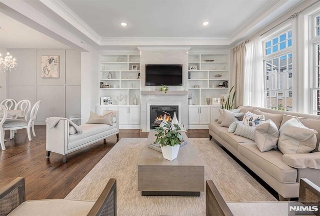 living area with a warm lit fireplace, ornamental molding, wood finished floors, built in shelves, and a notable chandelier