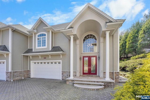 view of exterior entry featuring decorative driveway, stone siding, french doors, roof with shingles, and a garage