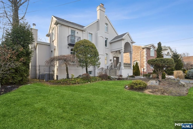 view of front of home featuring a balcony, fence, stucco siding, a chimney, and a front yard