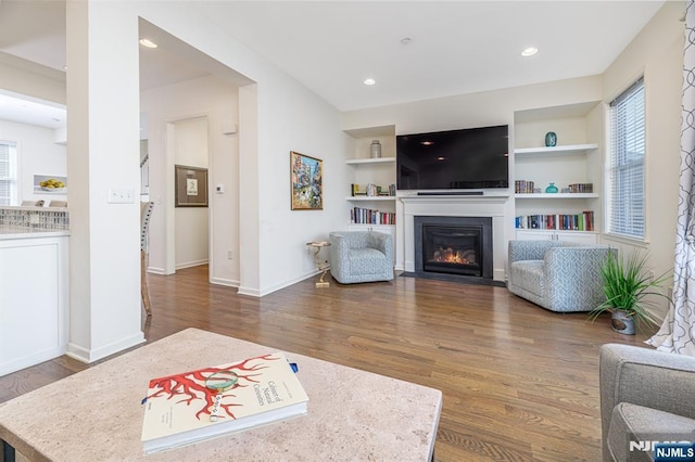 living room featuring built in shelves, wood finished floors, baseboards, a fireplace with flush hearth, and recessed lighting