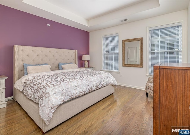 bedroom with a tray ceiling, wood finished floors, visible vents, and baseboards