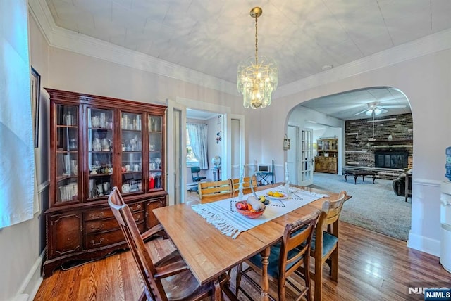 dining space featuring arched walkways, crown molding, a fireplace, wood finished floors, and ceiling fan with notable chandelier