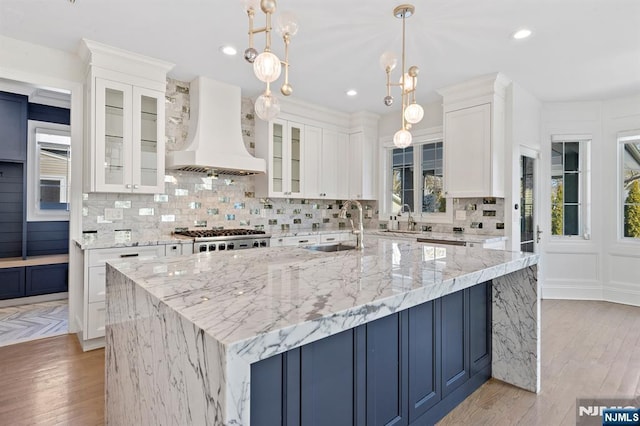 kitchen with a sink, white cabinetry, backsplash, custom exhaust hood, and a center island with sink