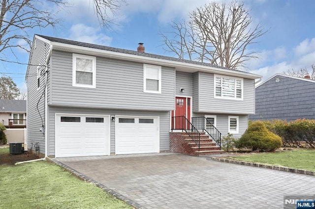 bi-level home featuring decorative driveway, a chimney, a front yard, a garage, and cooling unit