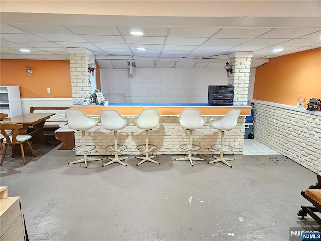 bar featuring a bar, a drop ceiling, concrete flooring, and wainscoting