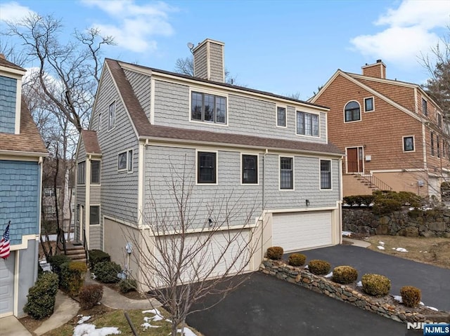 rear view of house with a garage, driveway, a chimney, and roof with shingles