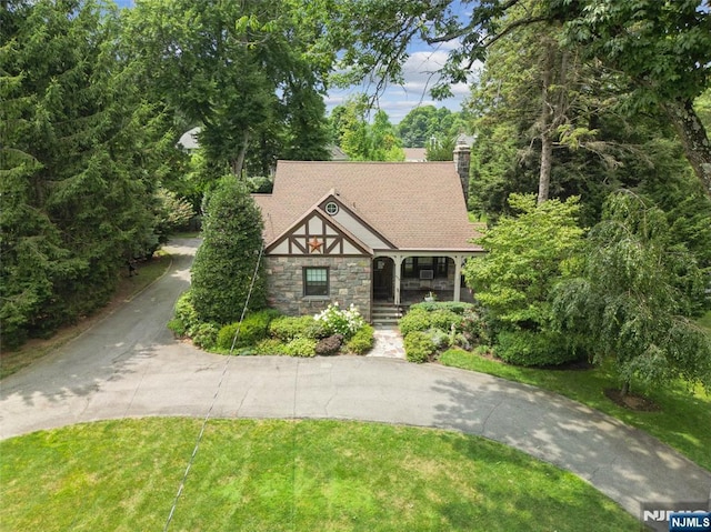 tudor-style house featuring a porch, a front yard, a chimney, stone siding, and driveway