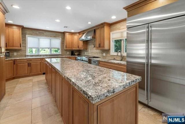 kitchen with stainless steel built in fridge, a kitchen island, a sink, brown cabinets, and wall chimney exhaust hood