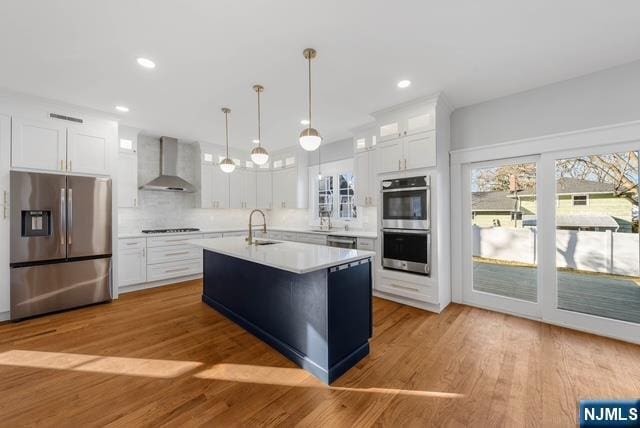 kitchen featuring light countertops, appliances with stainless steel finishes, white cabinets, a sink, and wall chimney range hood