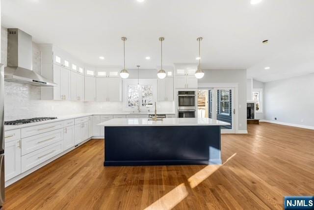 kitchen featuring light countertops, glass insert cabinets, stainless steel double oven, a kitchen island, and wall chimney exhaust hood