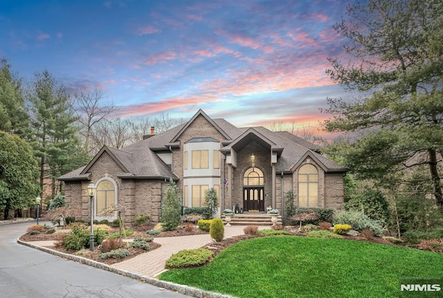 french country inspired facade featuring a lawn, decorative driveway, a chimney, and brick siding