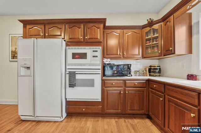 kitchen featuring white appliances, tasteful backsplash, brown cabinetry, glass insert cabinets, and light wood-type flooring