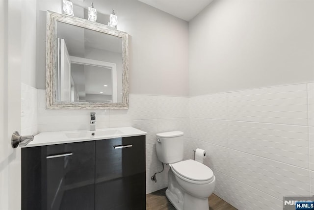 half bathroom featuring a wainscoted wall, tile walls, toilet, vanity, and wood finished floors