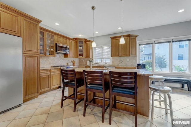 kitchen featuring brown cabinetry, light stone countertops, an island with sink, appliances with stainless steel finishes, and a kitchen bar