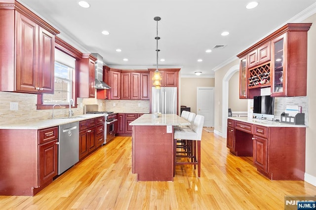 kitchen with wall chimney range hood, reddish brown cabinets, appliances with stainless steel finishes, and a sink