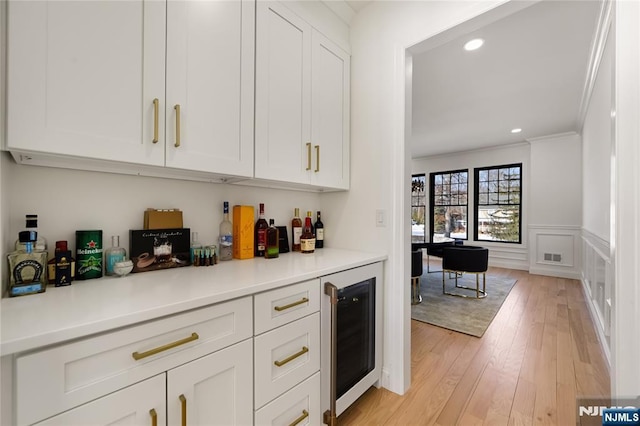 bar with beverage cooler, wainscoting, crown molding, light wood-style floors, and a decorative wall