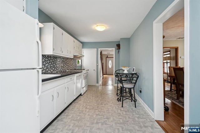 kitchen featuring tasteful backsplash, dark countertops, white cabinets, white appliances, and baseboards