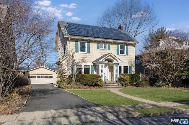 colonial-style house featuring a front yard, roof mounted solar panels, a chimney, and an outbuilding