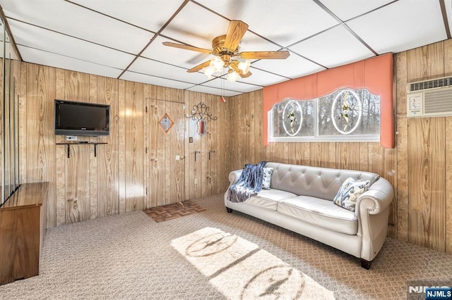 living area featuring a paneled ceiling, ceiling fan, a wall mounted air conditioner, carpet floors, and wood walls