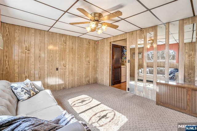 living area featuring carpet floors, a paneled ceiling, wooden walls, and a ceiling fan